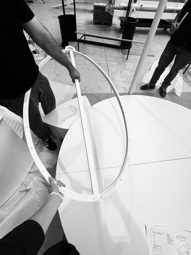 Dettaglio in bianco e nero della fase di assemblaggio del prototipo Moonglow: mani del team al lavoro su un elemento circolare strutturale, con componenti tecnici e disegni esecutivi visibili durante il processo produttivo