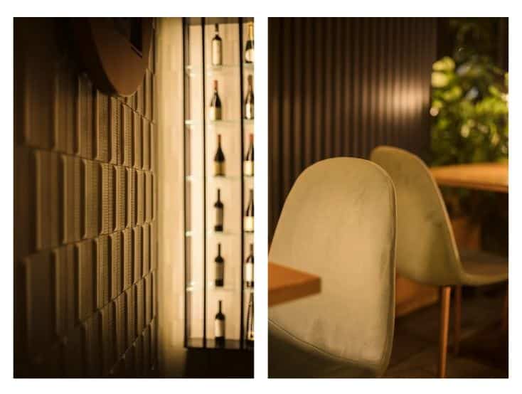Immagine a sinistra: Dettaglio materico di una parete rivestita con elementi tridimensionali geometrici, illuminata da una luce calda che ne esalta profondità e texture. Sullo sfondo, una vetrina verticale con bottiglie retroilluminate crea un elegante contrasto tra superficie opaca e luce diffusa. Immagine a destra: Primo piano di sedute imbottite dai toni chiari e morbidi, abbinate a tavoli con piano in legno. Sullo sfondo, una parete a doghe verticali e una presenza vegetale contribuiscono a creare un’atmosfera accogliente e raffinata, giocata su luci soffuse e materiali naturali.