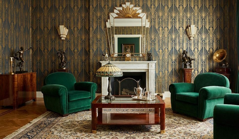 Elegante salotto in stile Art Déco anni ’20-’30 con poltrone in velluto verde smeraldo, camino in marmo bianco, carta da parati geometrica dorata e blu, tavolino in legno lucido e dettagli vintage raffinati. Atmosfera sofisticata e lussuosa tipica delle dimore d’epoca europee.