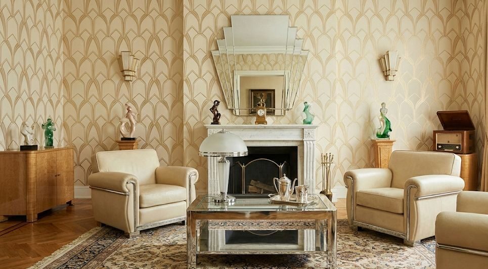 Raffinato salotto ispirato agli anni ’40 e ’50 con poltrone imbottite color crema, camino in marmo, specchio geometrico Art Déco e mobili in legno dalle linee eleganti. La carta da parati decorativa e gli accessori vintage creano un’atmosfera calda, sofisticata e senza tempo, tipica delle case di lusso del dopoguerra.