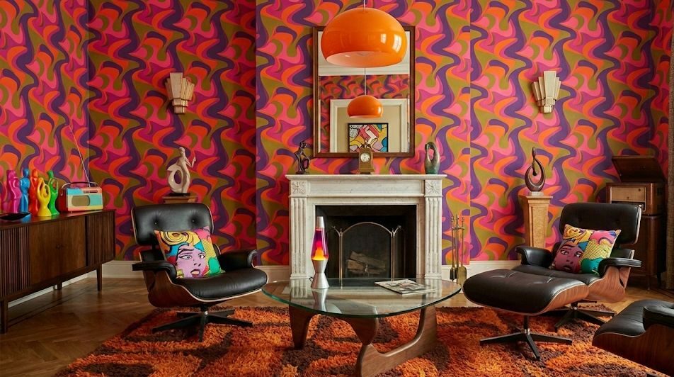 Vivace salotto in stile anni ’60 e ’70 con carta da parati psichedelica dai colori arancione, rosa e viola, poltrone lounge in pelle nera e legno curvato, lampada a sospensione rétro e tavolino in vetro. L’ambiente richiama il design pop e futurista dell’epoca, con dettagli vintage, forme morbide e atmosfera creativa e anticonvenzionale.