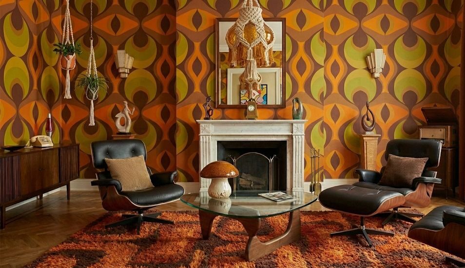 Salotto in perfetto stile anni ’60 e ’70 con carta da parati geometrica rétro nei toni arancio, marrone e verde oliva, poltrone lounge iconiche in pelle nera e legno curvato, tappeto shaggy e dettagli boho come macramè sospesi. L’ambiente esprime il carattere caldo, creativo e psichedelico tipico del design vintage di quell’epoca.