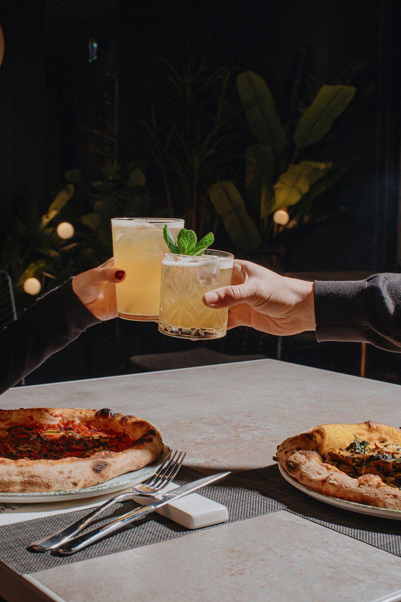 Scena conviviale in un ristorante, con due persone che brindano sollevando cocktail chiari con ghiaccio e foglie di menta. In primo piano, su un tavolo in pietra chiara, sono servite due pizze artigianali dai bordi alti e ben dorati, accompagnate da posate disposte con cura. Lo sfondo è scuro e suggestivo, arricchito da piante e luci soffuse che creano un’atmosfera intima ed elegante.