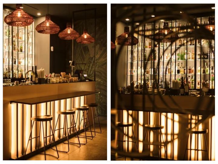 Immagine di progettazione interior design horeca a sinistra: Bancone bar dal design contemporaneo, caratterizzato da una base retroilluminata che crea un effetto scenografico caldo e avvolgente. Sopra, una fila di lampade a sospensione in fibra naturale diffonde una luce soffusa, valorizzando il piano del banco e gli sgabelli alti con struttura metallica e seduta minimale. Sullo sfondo, una parete attrezzata con bottiglie esposte completa l’ambiente, rendendolo accogliente e curato nei dettagli. Immagine a destra: Vista più ravvicinata e suggestiva del bancone, con un gioco di luci e ombre creato da elementi decorativi in primo piano. La retroilluminazione del banco enfatizza le geometrie verticali, mentre la parete con bottiglie illuminate sullo sfondo aggiunge profondità e atmosfera. L’insieme restituisce uno spazio intimo, raffinato e visivamente dinamico.