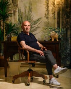 Foto di Nicola Lucio Ruscino, imprenditore materano doc, seduto in uno studio elegante con arredi in legno e sfondo botanico, in un’atmosfera raffinata e professionale.