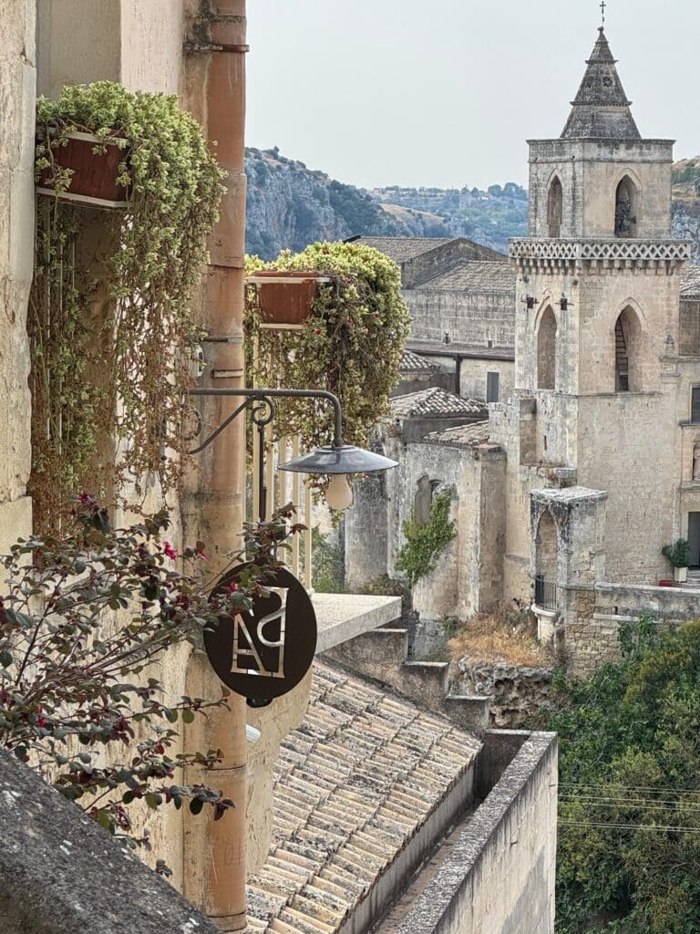 Vista panoramica dai Sassi di Matera con balcone fiorito e campanile storico sullo sfondo, scattata dall’Hotel Sant’Angelo, struttura di charme immersa nel paesaggio unico della città patrimonio UNESCO.