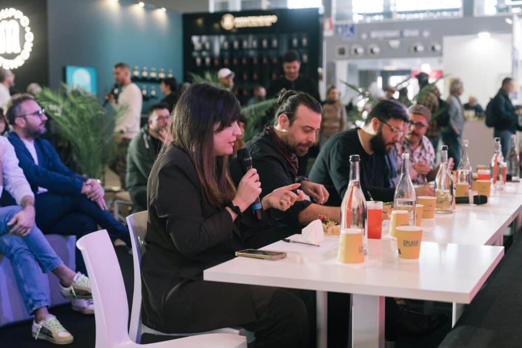 Silvia Orlandi interviene con microfono al tavolo dei giudici durante la Pizza al Padellino & Cocktail Competition allo Splash Hospitality Expo di Bari, mentre assaggia e valuta le proposte insieme agli altri giurati. Sul tavolo sono presenti drink, bottiglie e piatti in degustazione, in un contesto vivace e professionale con pubblico e stand sullo sfondo.